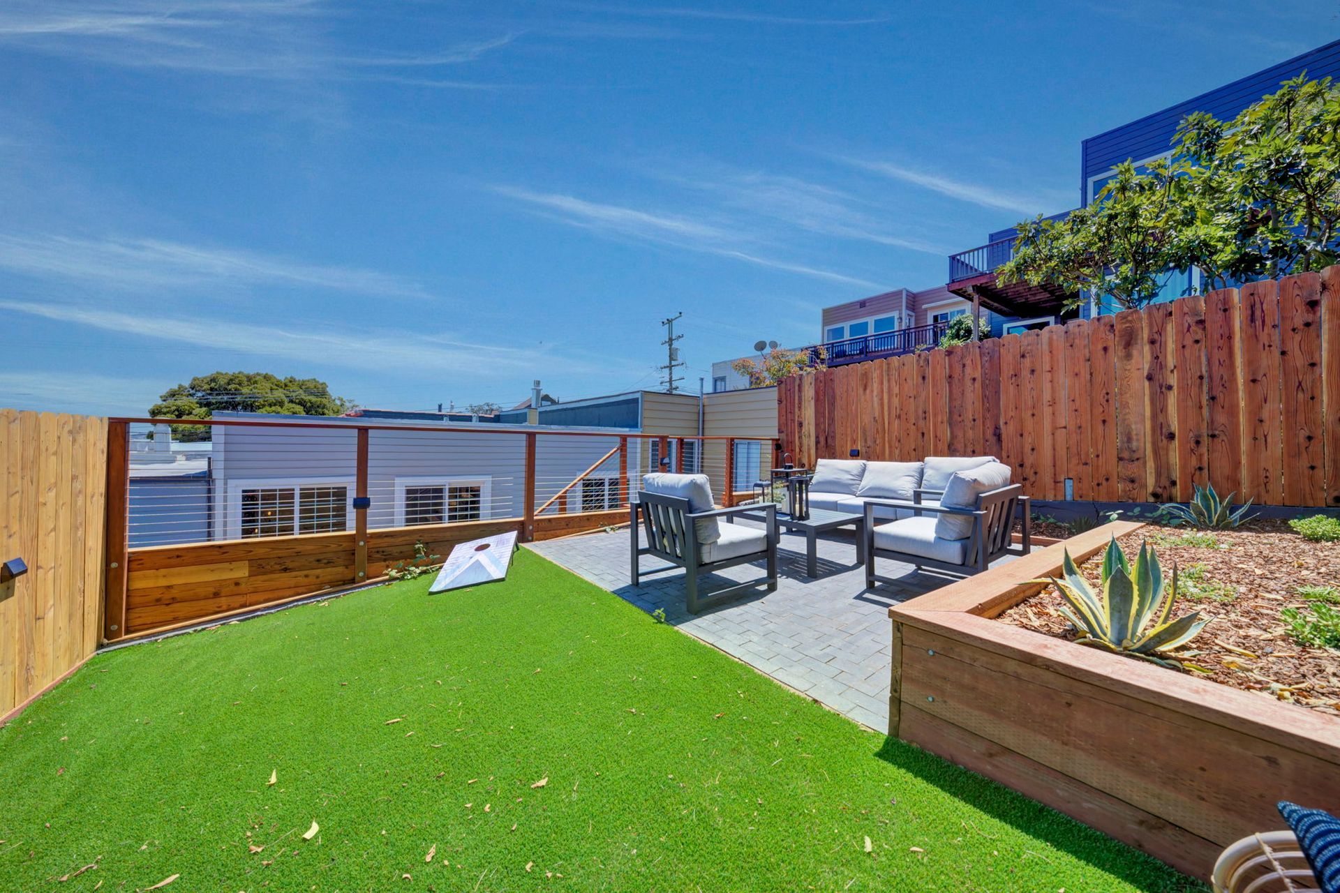 Sunny backyard patio with lawn, outdoor seating, raised garden bed, and wooden fence