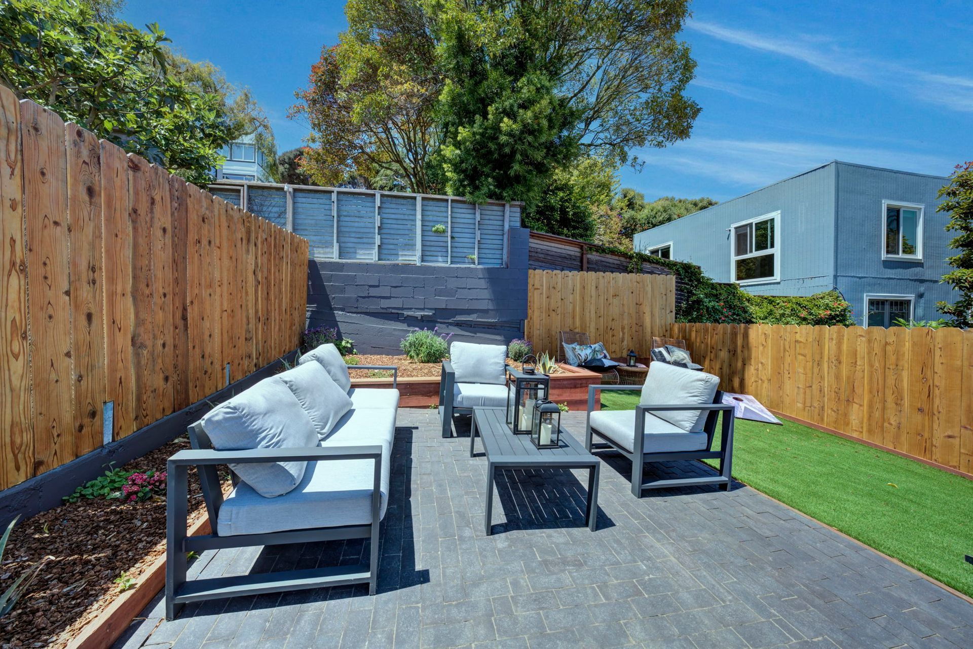 Modern backyard patio with white outdoor seating, small table, wooden fence, and artificial grass