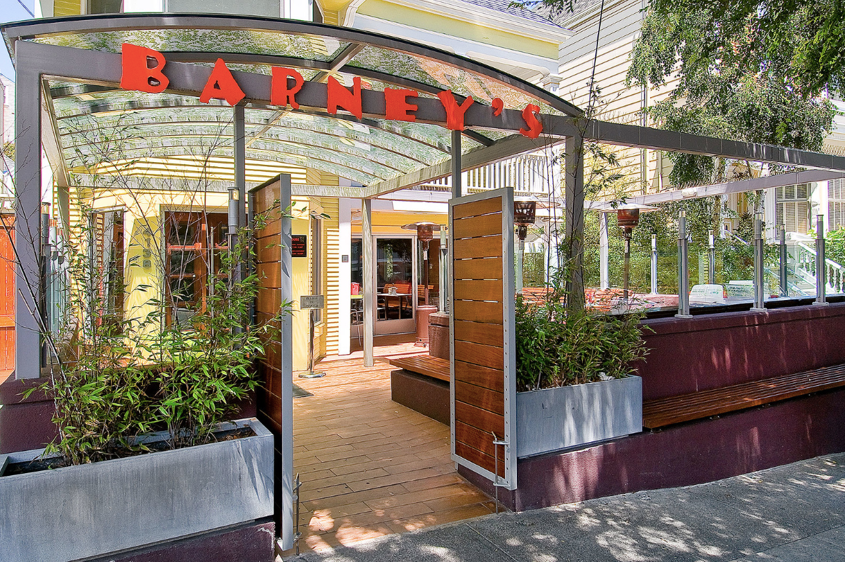 Bakery storefront with red sign, glass doors, and outdoor planters on a sunny street