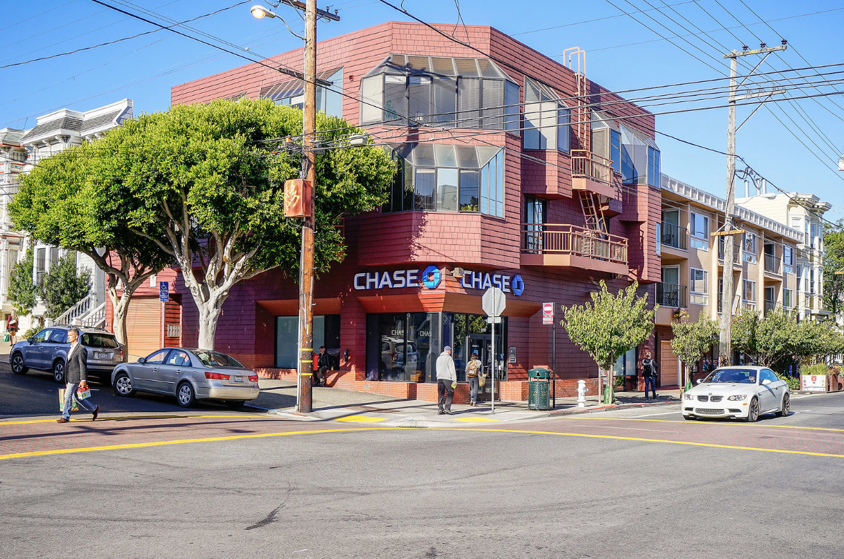 Corner street view of a red brick Chase bank building with trees, parked cars, and overhead cables.