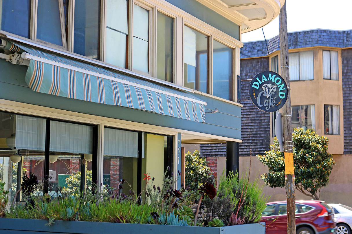 Street-level view of a cafe with blue awning, large windows, potted plants, and a round hanging sign.