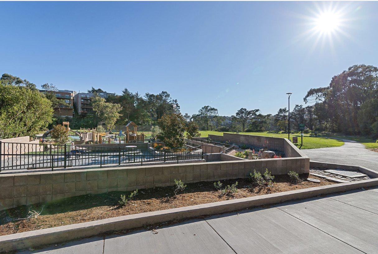 Sunny outdoor park with benches, low walls, and trees along a paved road