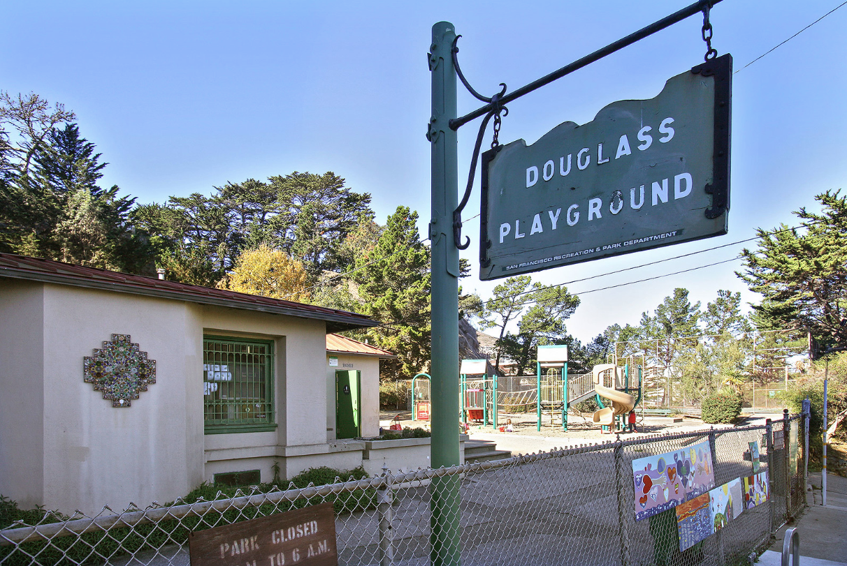 Entrance sign for Douglass Playground beside a small building and fenced play area on a sunny day