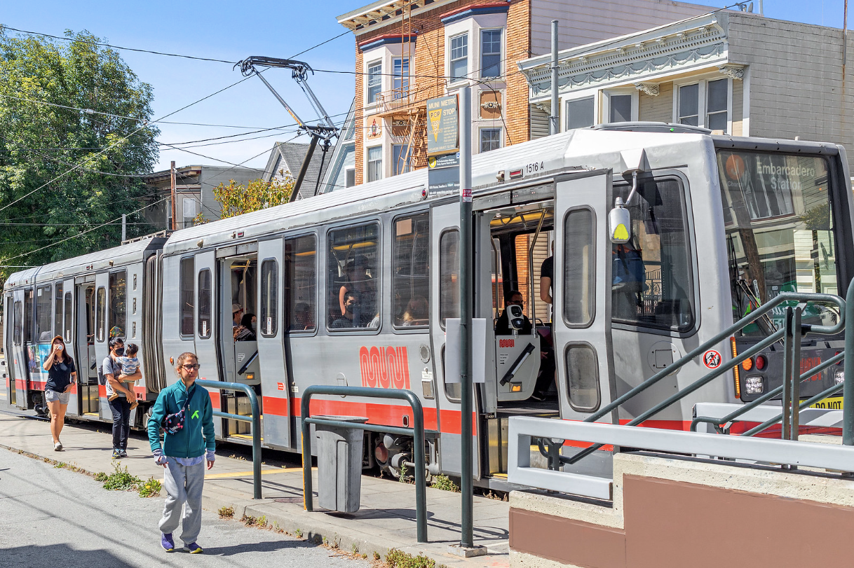 Modern streetcar stopped at a platform with passengers and a pedestrian nearby, in front of buildings and trees