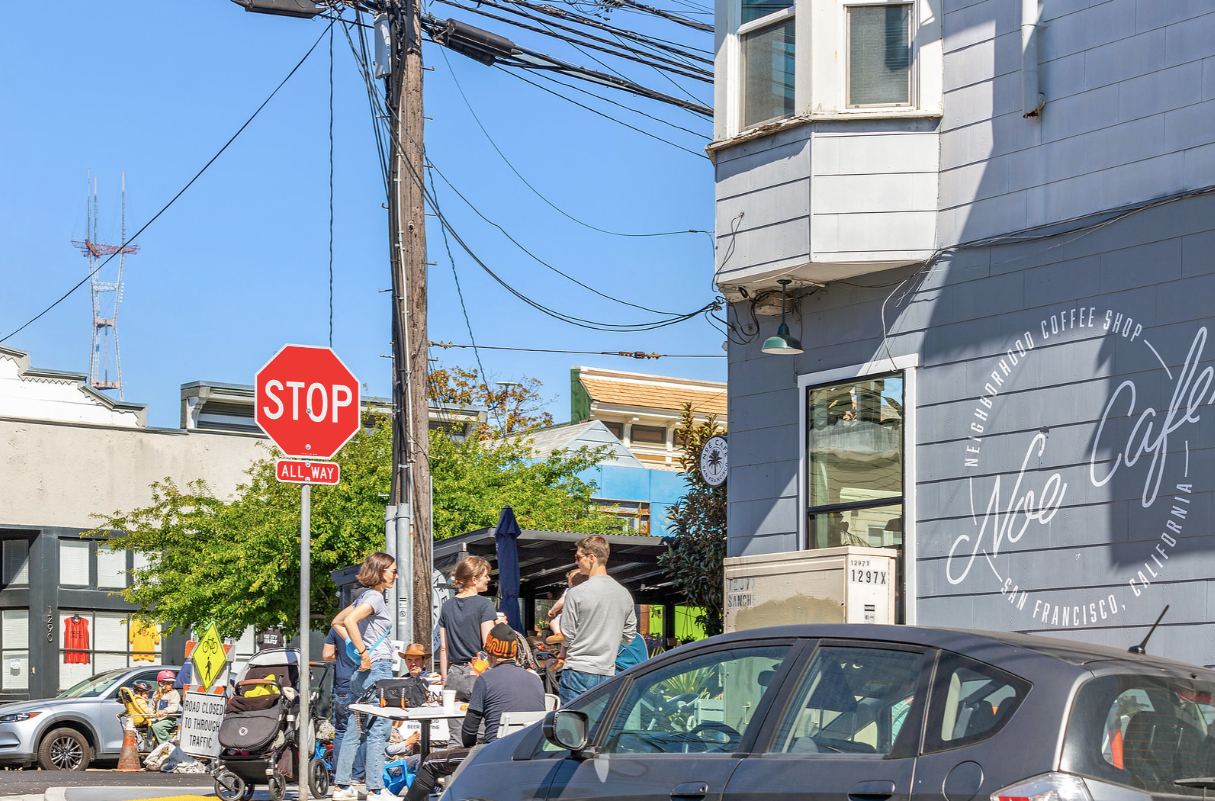 Street café scene with parked cars, a stop sign, and people sitting outdoors on a sunny corner