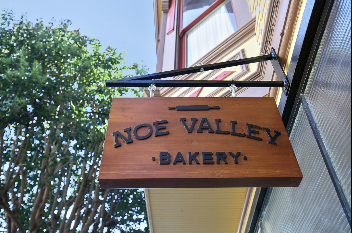Wooden hanging sign reading “Noe Valley Bakery” outside a storefront, with trees in the background