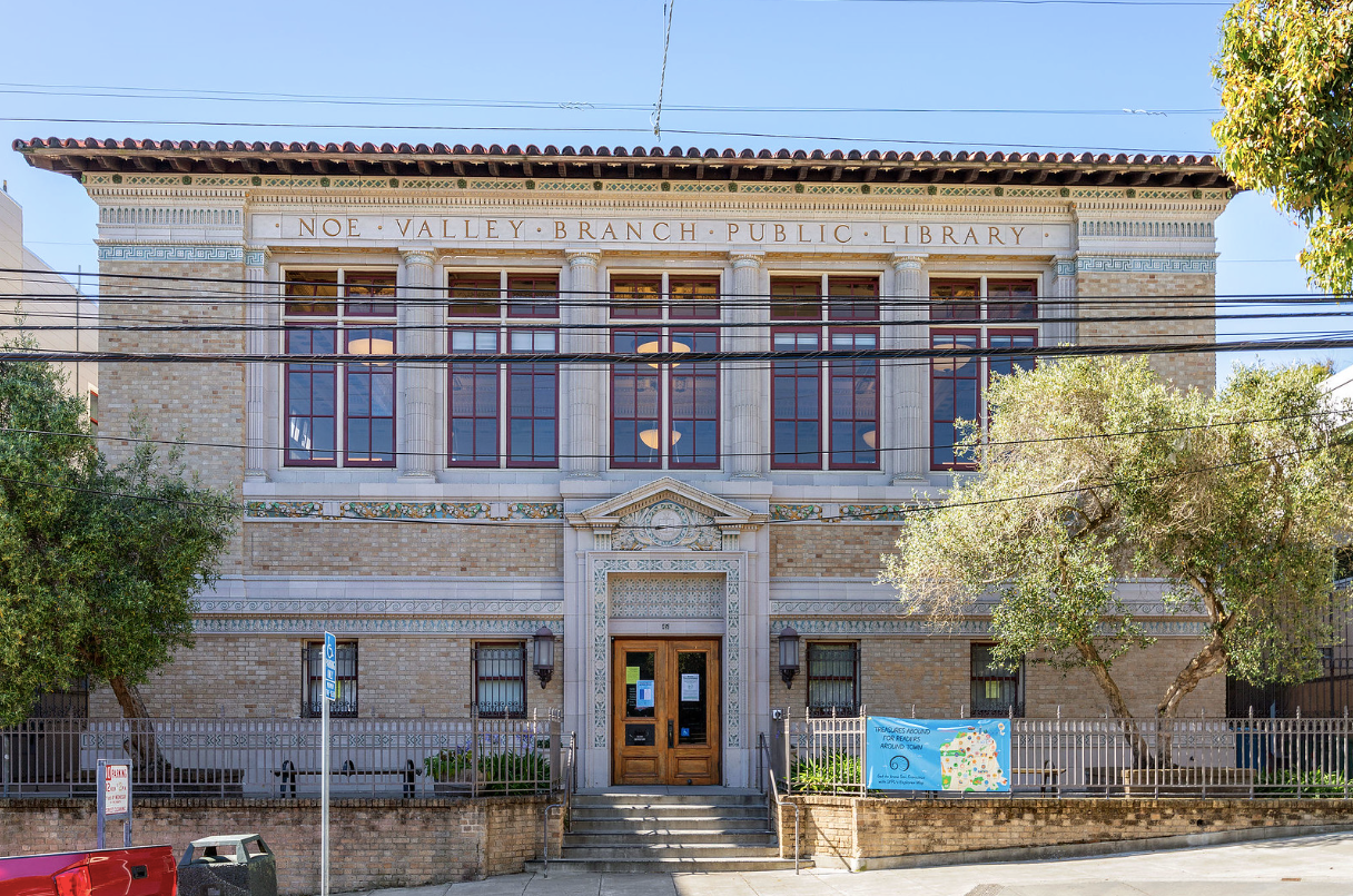 Historic two-story stone building with central wooden doors, tall windows, and trees lining the street
