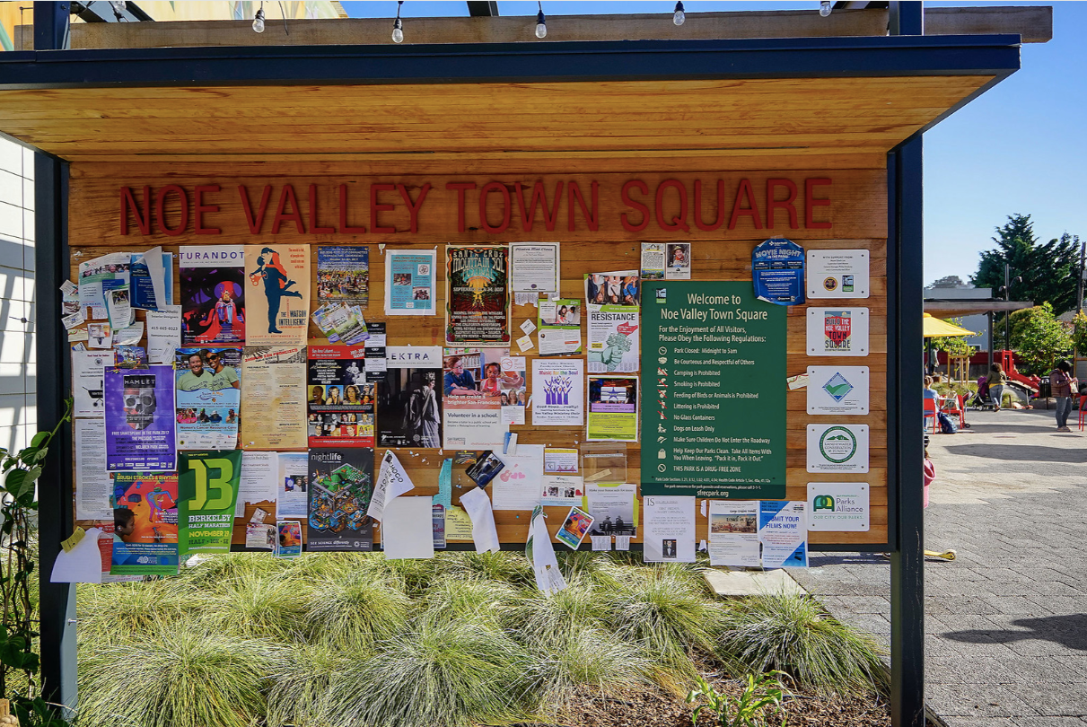 Noc Valley Town Square bulletin board with flyers, notices, and a green information panel outdoors under a roof.