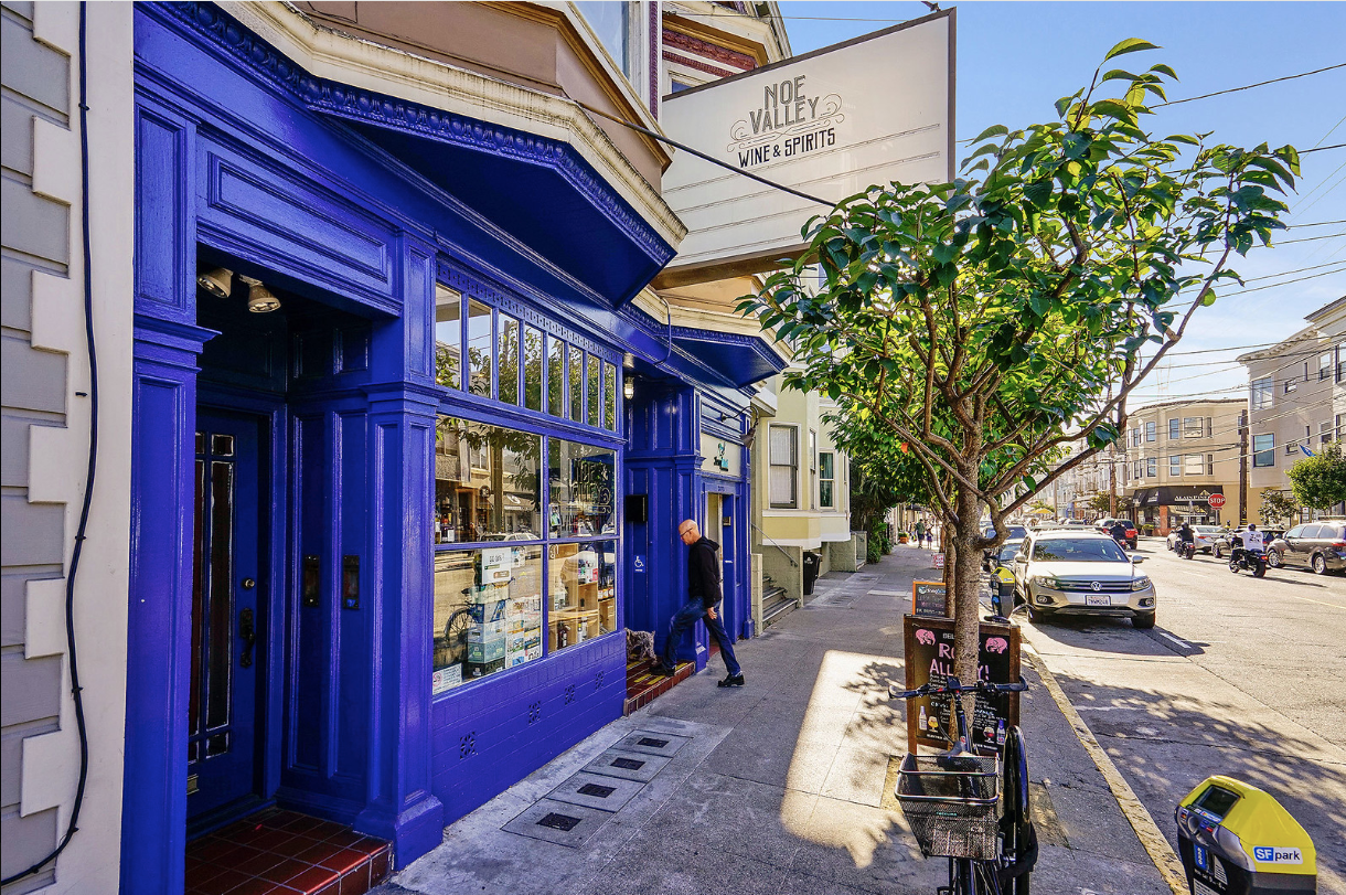 Bright blue storefront on a sunny city street with trees, parked cars, and buildings lining the sidewalk