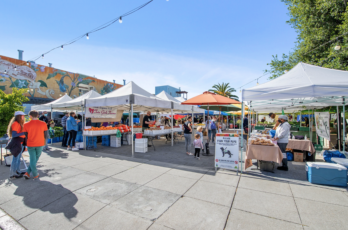 Outdoor market with white tents, vendors, and shoppers on a sunny street.