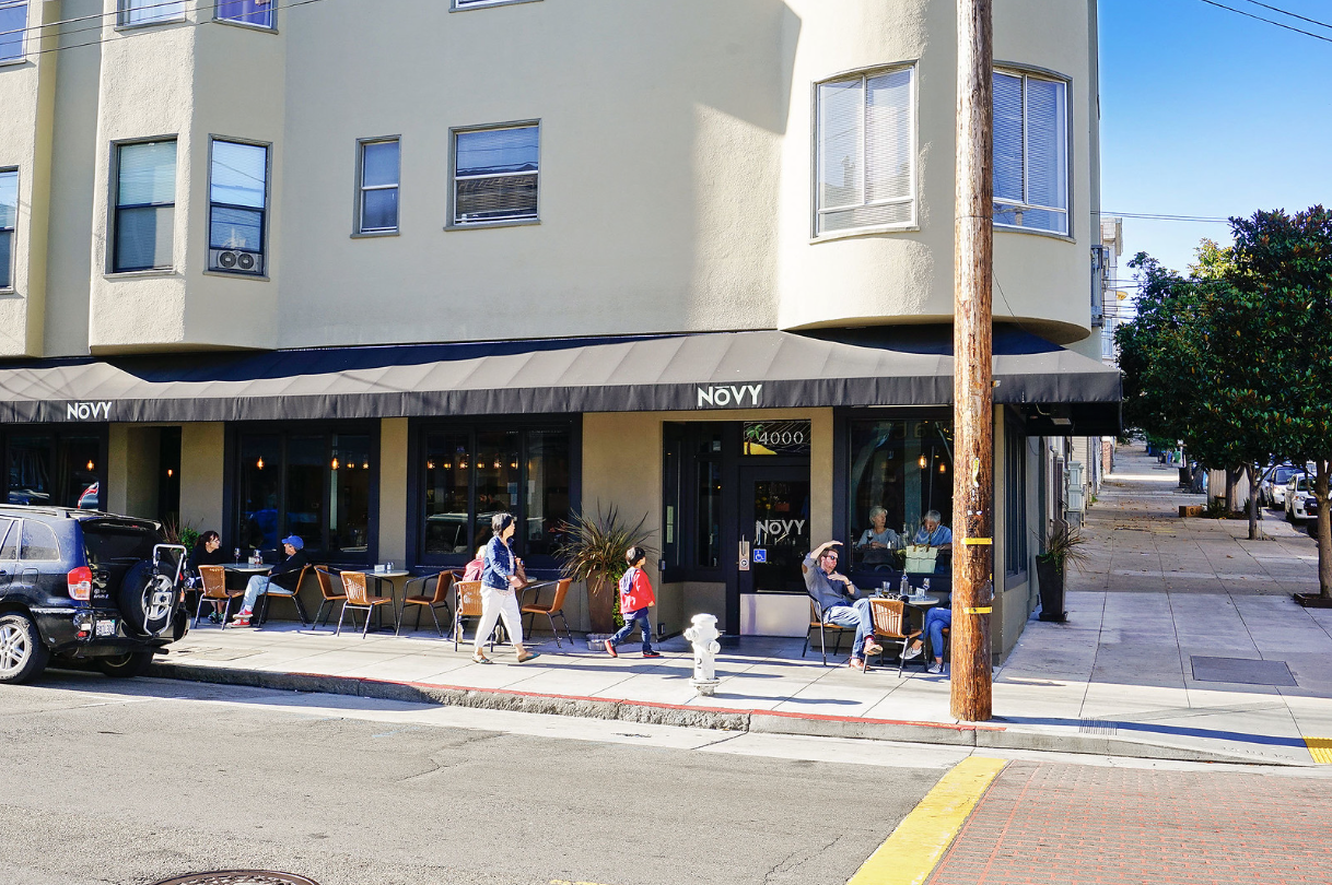 Outdoor café seating along a sunny street corner under a beige building with awning and pedestrians nearby