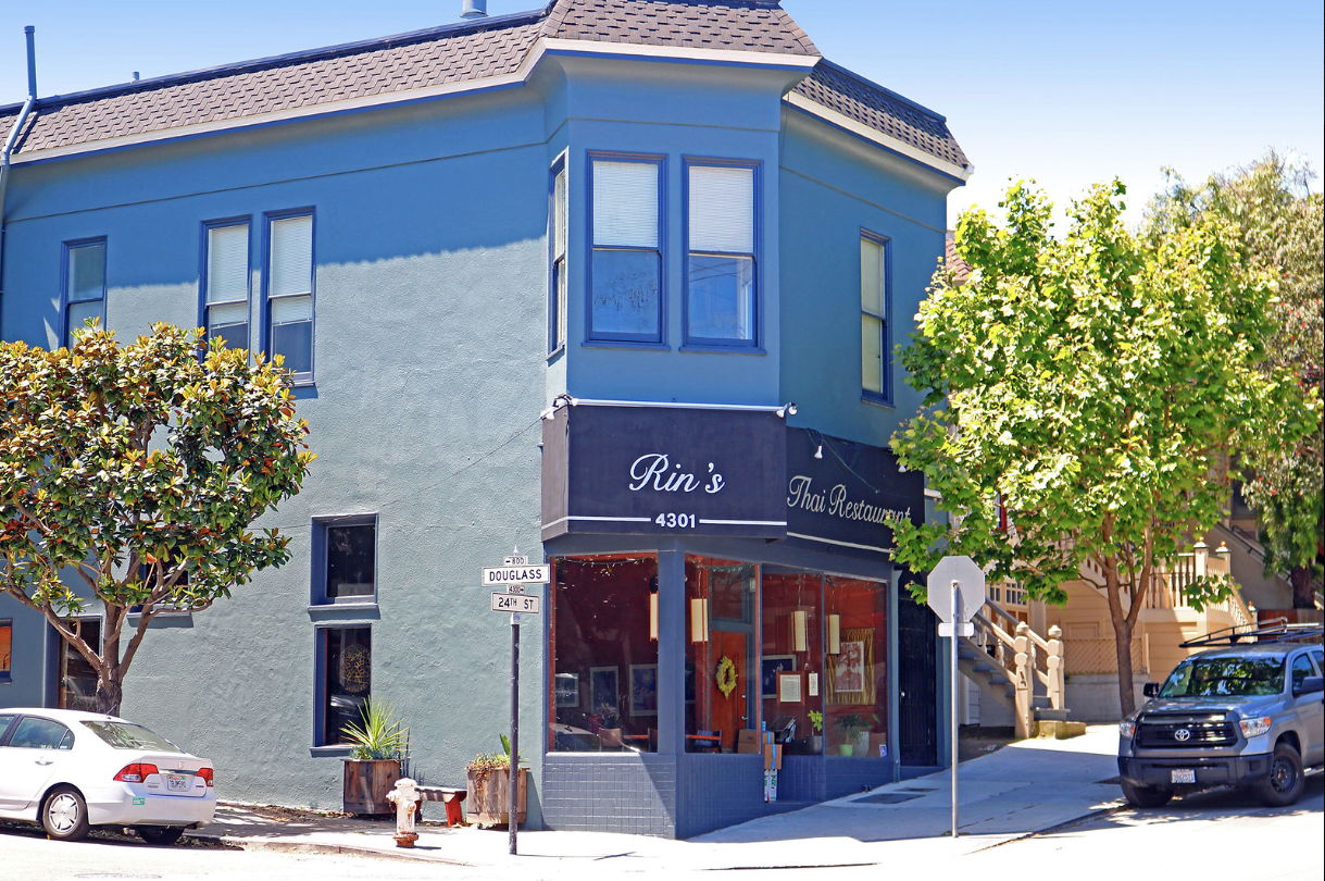 Corner storefront with blue facade, red awning, and trees on a sunny street.