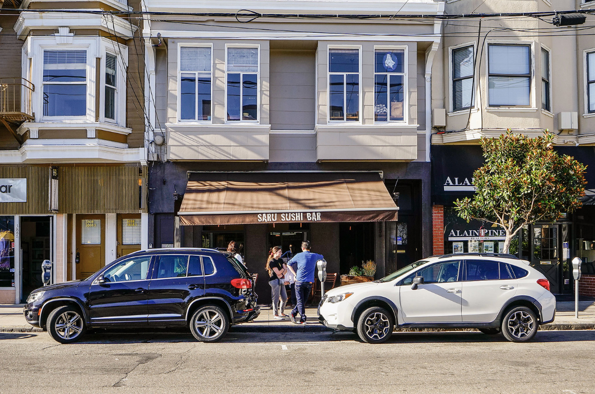 Two SUVs parked in front of storefronts, with people standing on the sidewalk between them