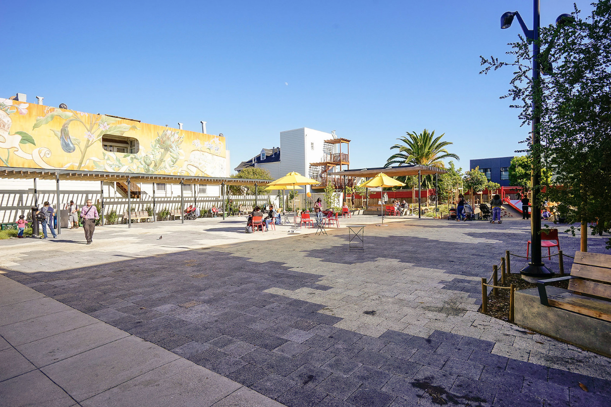 Sunny outdoor plaza with people walking, red umbrellas, and a yellow mural along the buildings