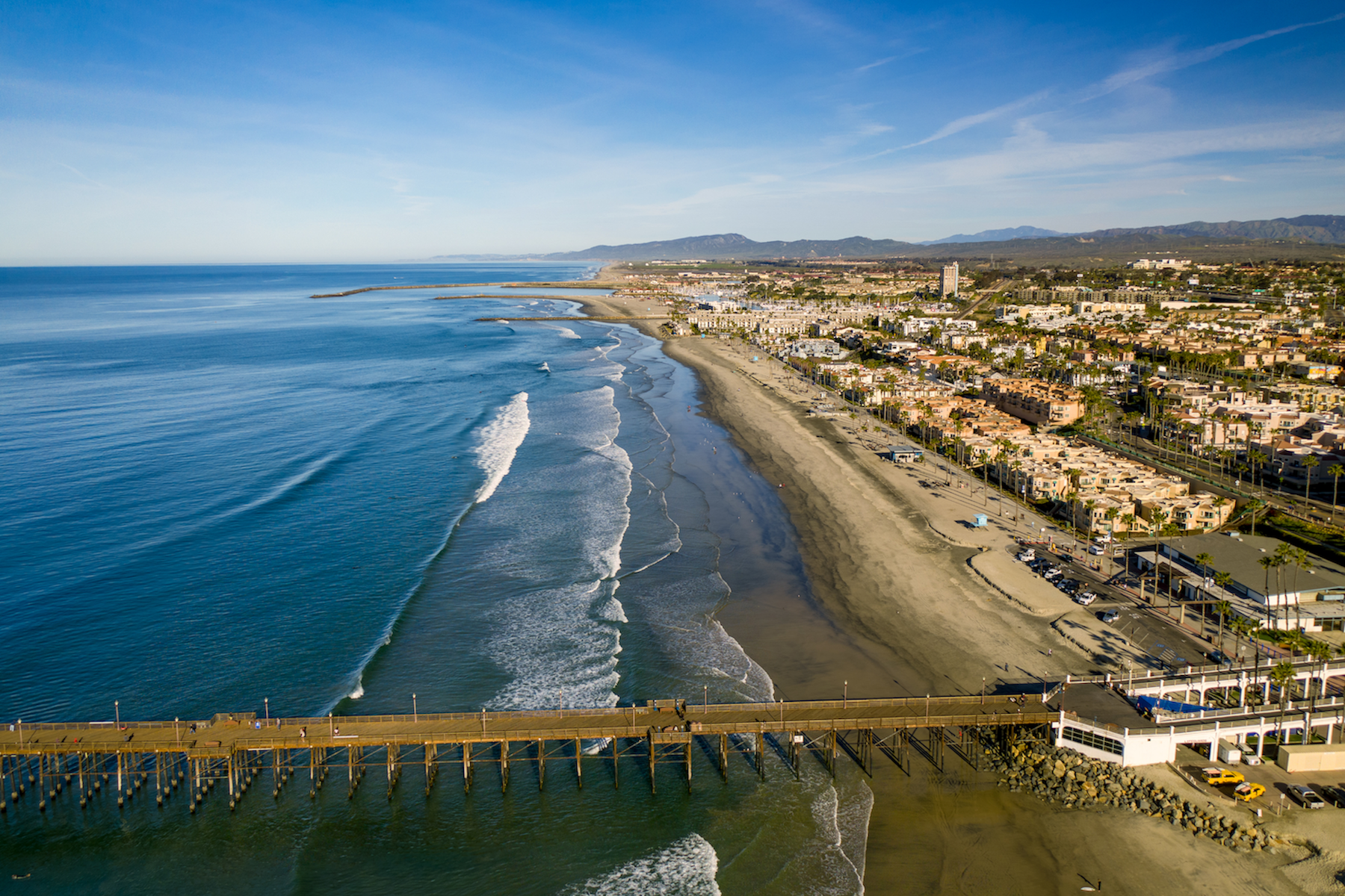 Aerial view of a sandy beach with a pier, ocean waves, and a coastal town along the shore.