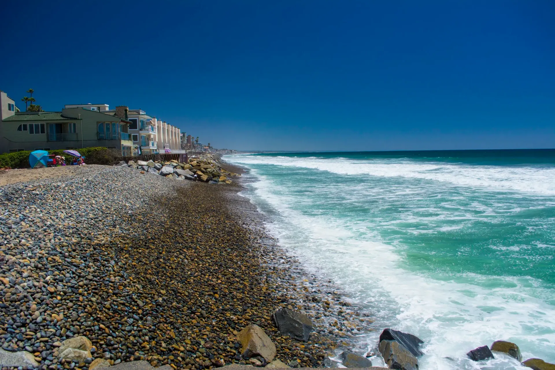 Rocky beach with white surf and blue ocean beside seaside buildings under a clear sky
