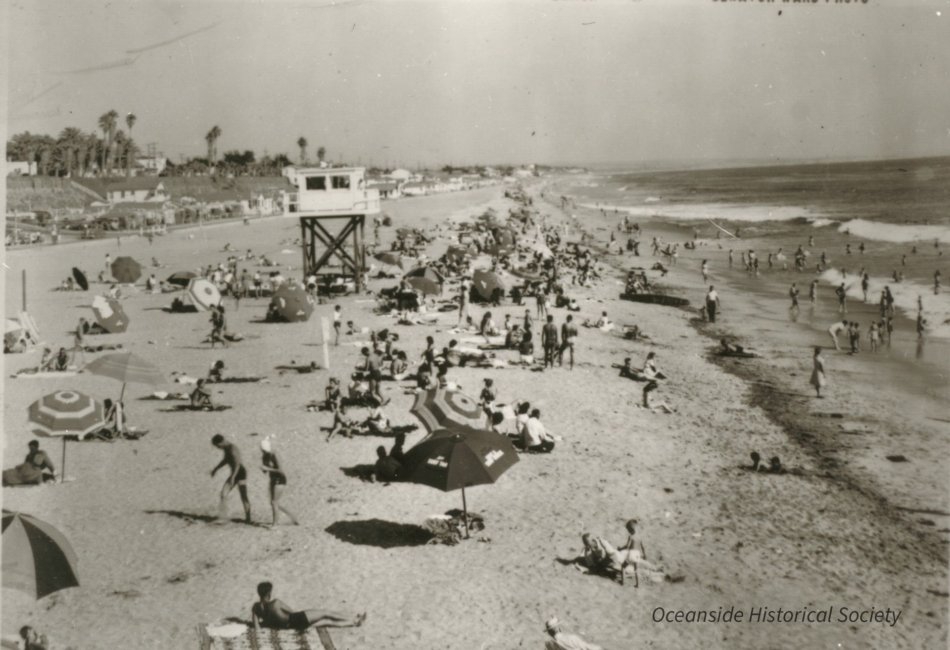 Crowded beach with umbrellas, swimmers, and a lifeguard tower along the shoreline
