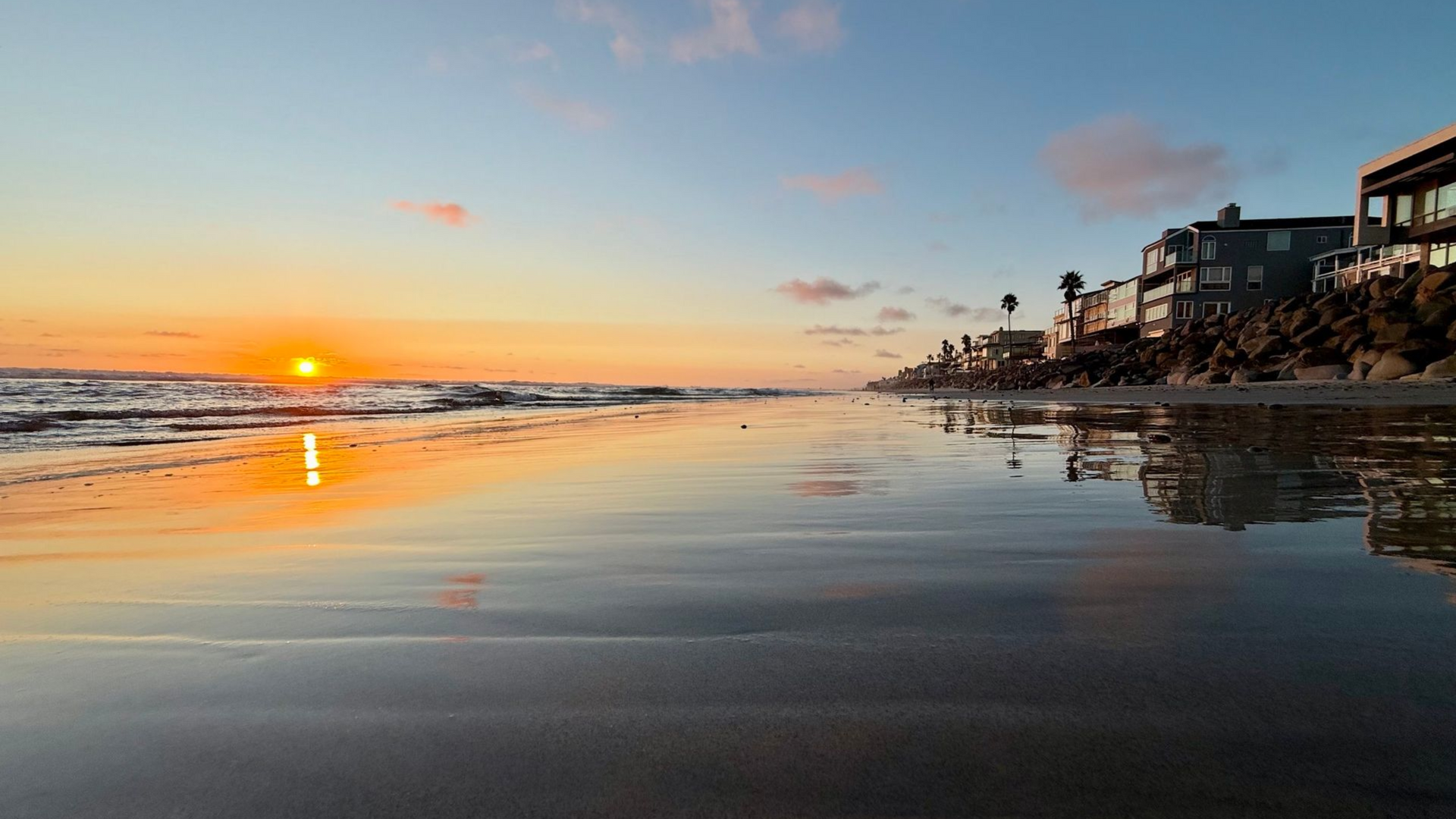 Sunset over a calm beach with wet sand reflecting orange sky and shoreline houses on the right