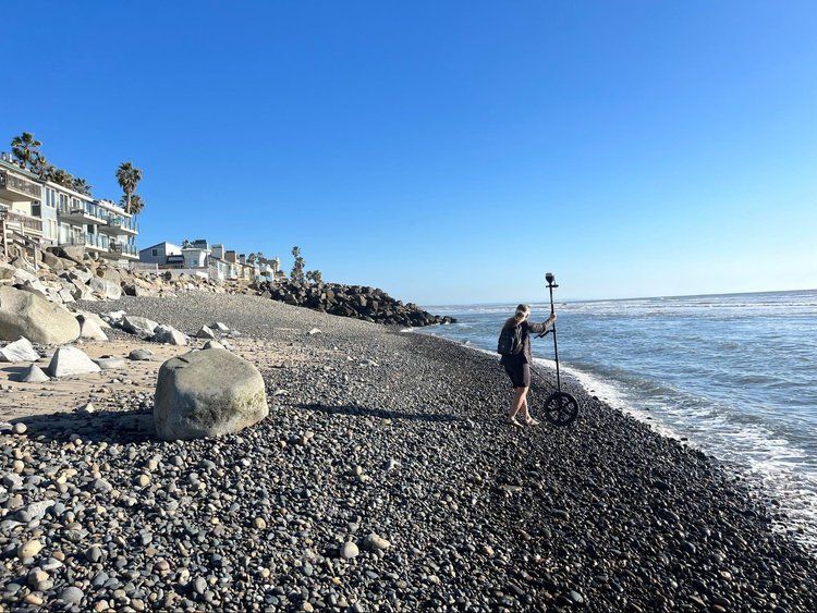 Person standing on a rocky beach beside the ocean, holding a fishing rod under a clear blue sky