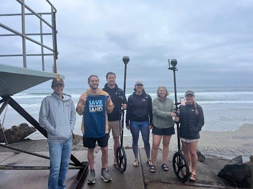 Group posing with e-standup paddleboards on a beach under cloudy skies