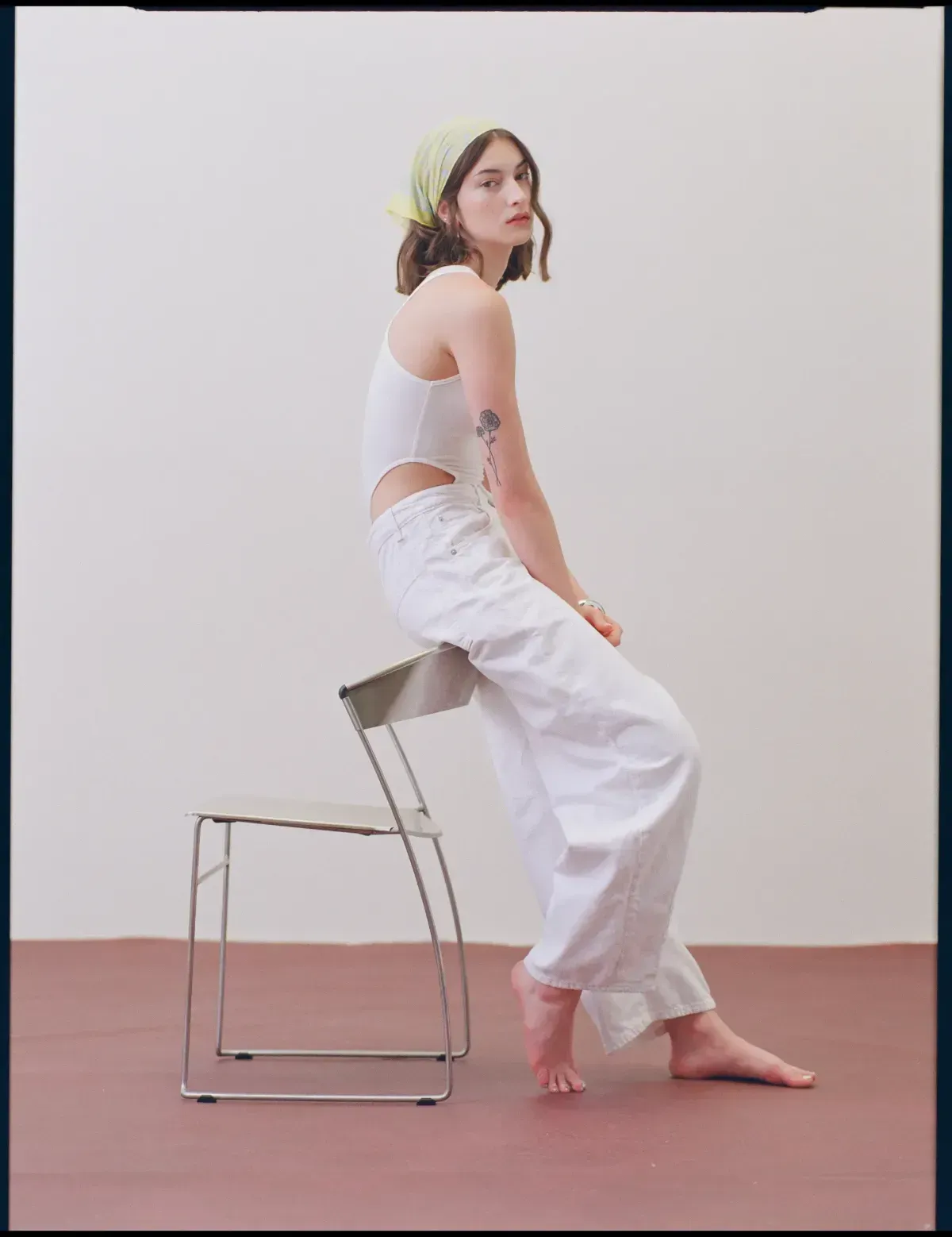 Person seated on a metal chair in a white top and pants against a plain light wall.