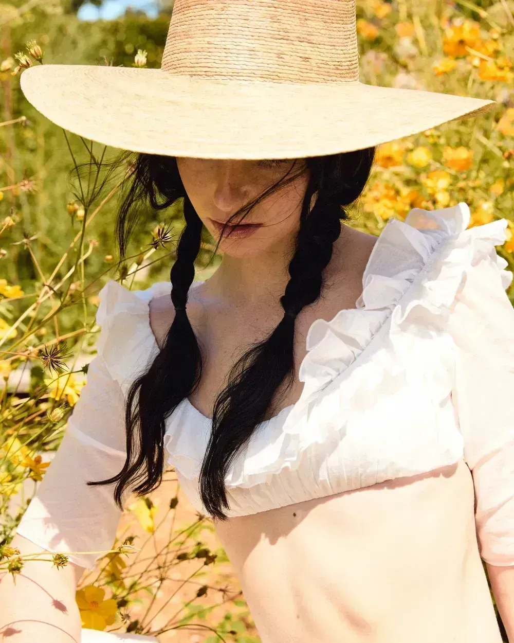 Woman in a straw hat and white ruffled top standing in a sunny field of yellow flowers.