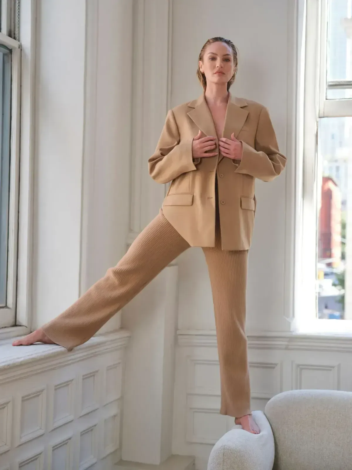 Person in beige suit posing by a tall window, one leg lifted on a ledge.