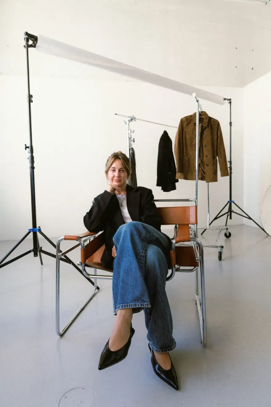 Woman sitting on a brown chair beside a wall, wearing a dark blue blouse and jeans.