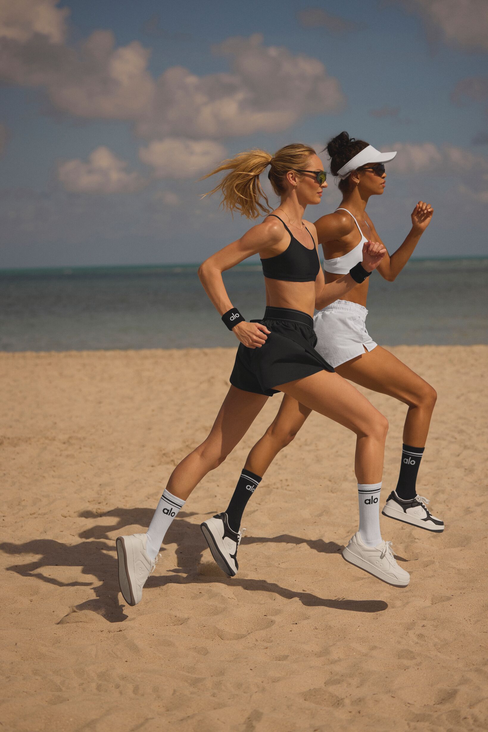 Two runners in sportswear jogging side by side on a sandy beach under a blue sky.