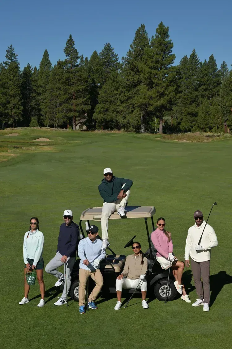 Golf group posing on a green with a man standing on a bench in the center, trees in the background