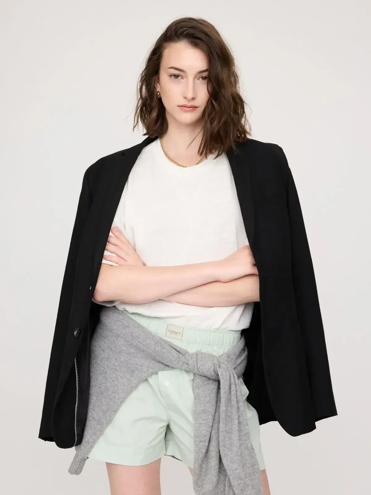 Person standing with arms crossed, wearing a black jacket over a white top and light shorts on a plain background
