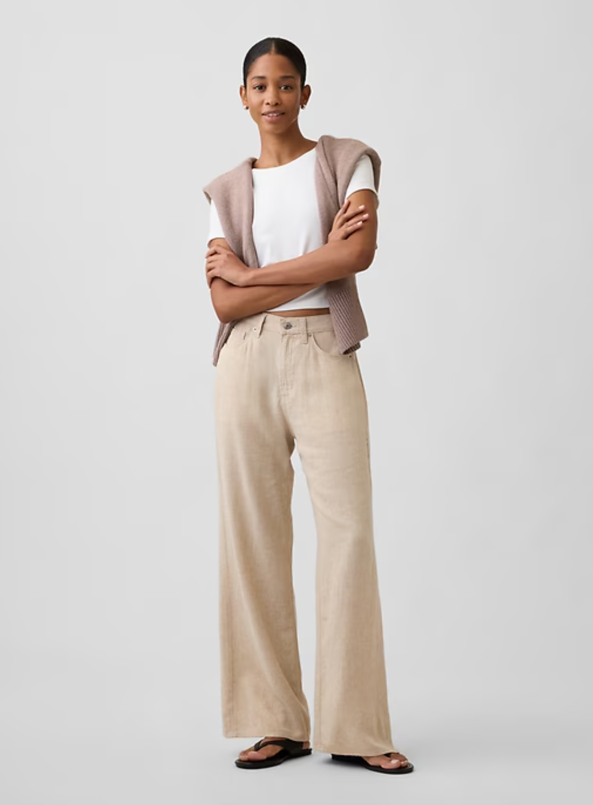 Person standing with arms crossed, wearing a beige outfit and black sandals on a plain light background