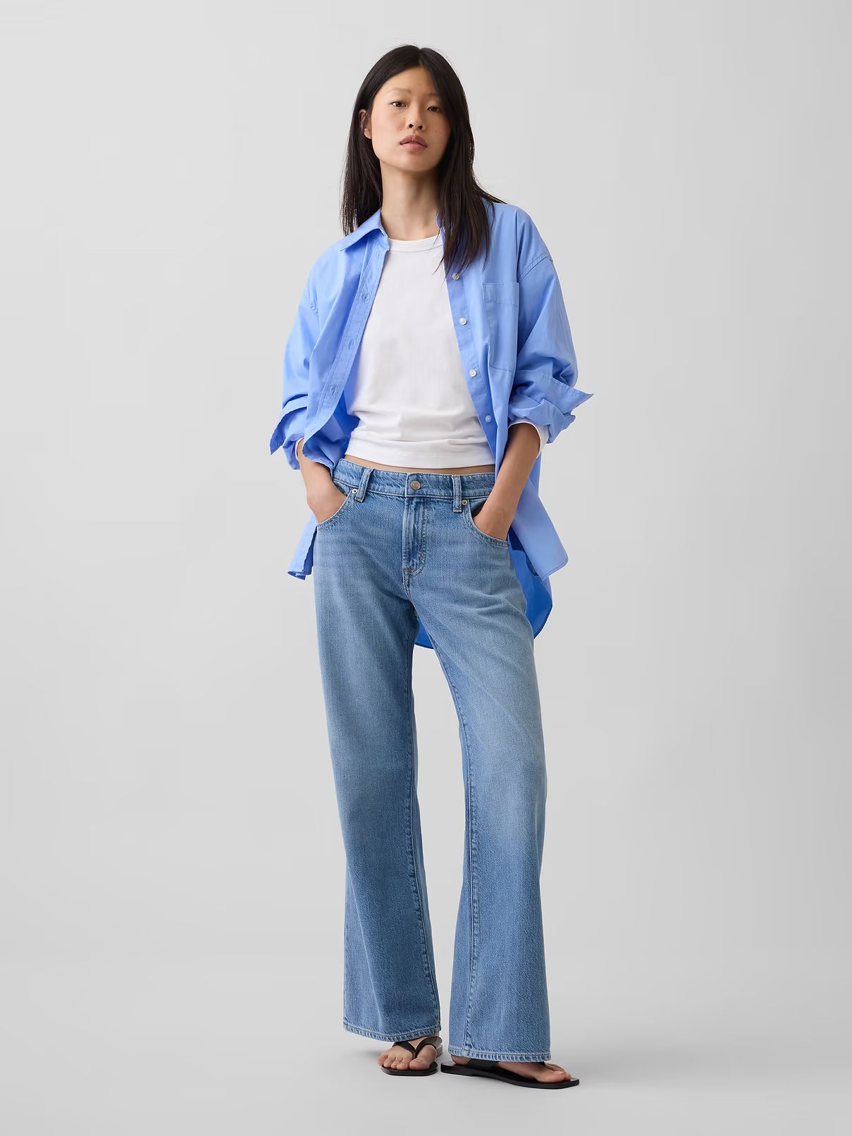 Woman in a blue shirt and jeans standing with hands in pockets on a plain background
