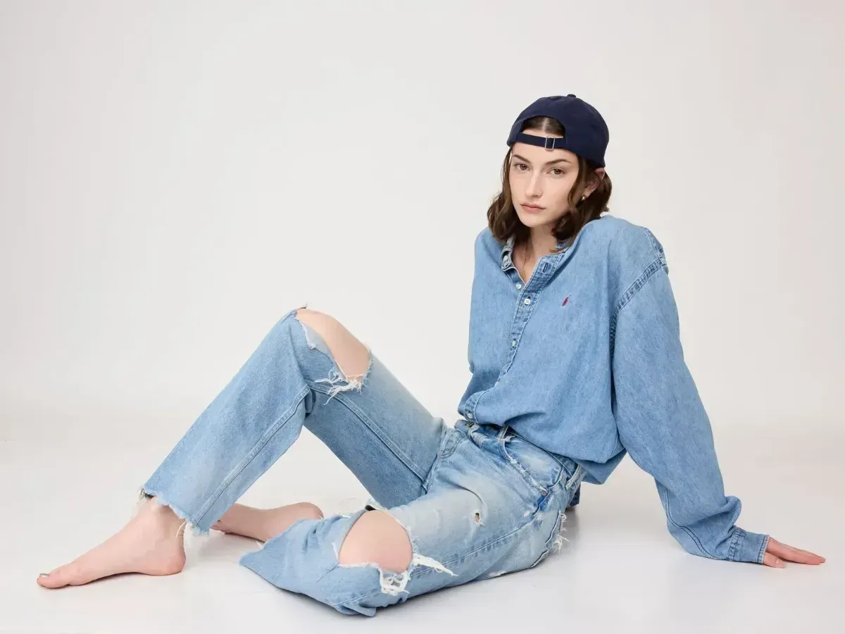 Woman in a blue outfit and backward cap posing seated against a white background.
