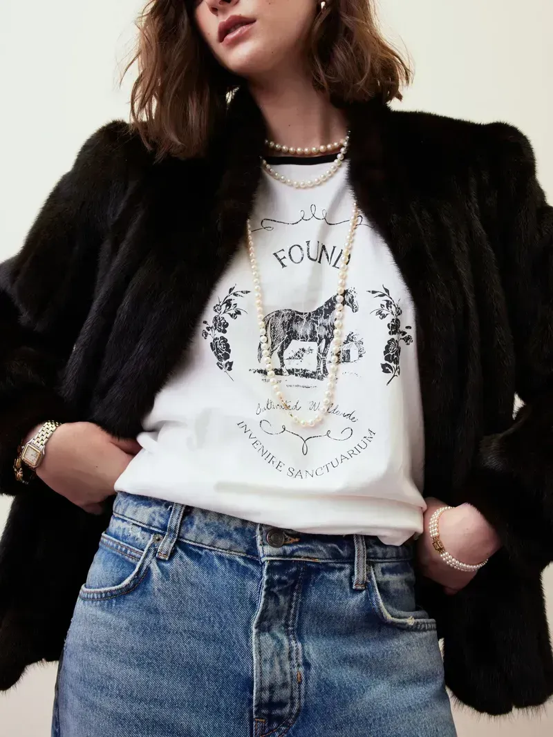 Person in black jacket and blue jeans wearing a white graphic T-shirt and layered necklaces.