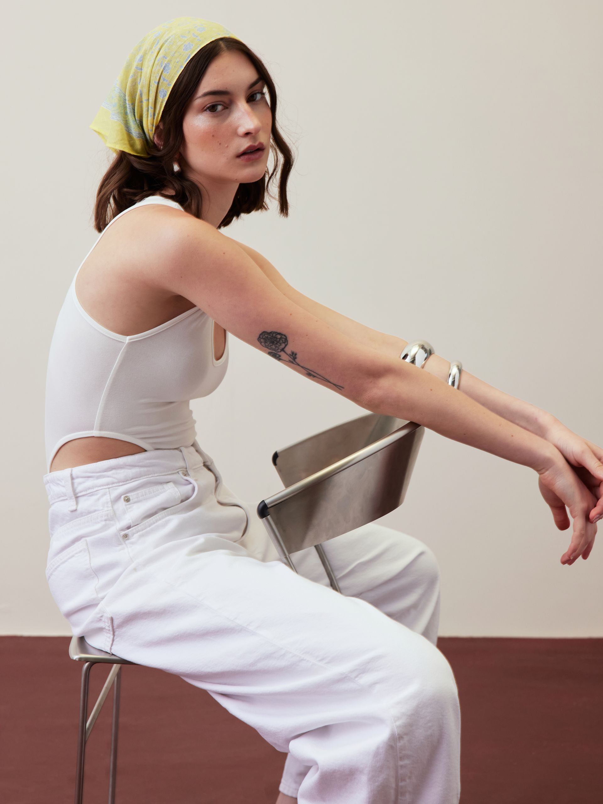 Person seated sideways on a metal chair, wearing a yellow headscarf, white top, and white pants against a plain wall