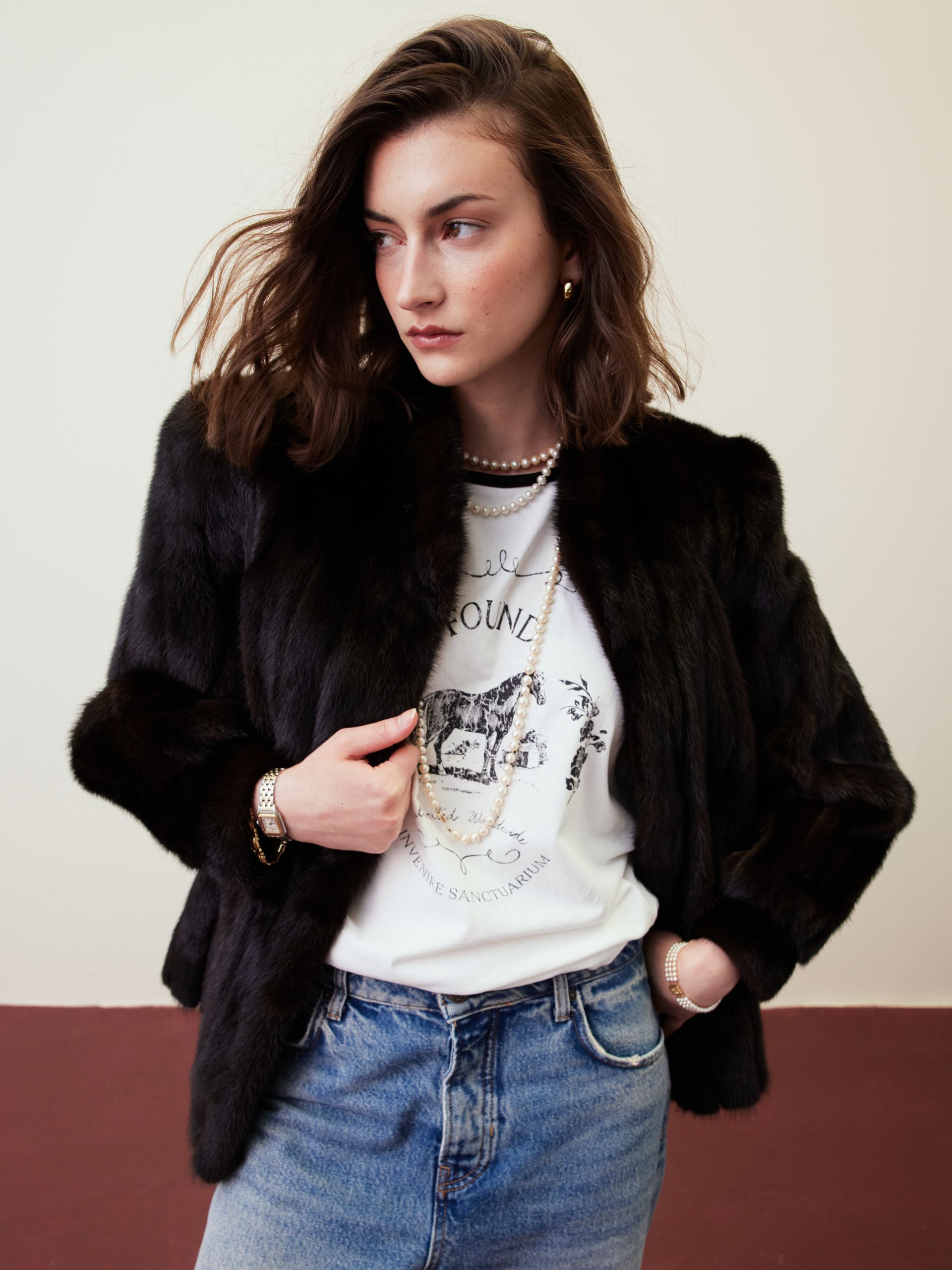 Person in a black fur jacket and jeans posing against a light wall.
