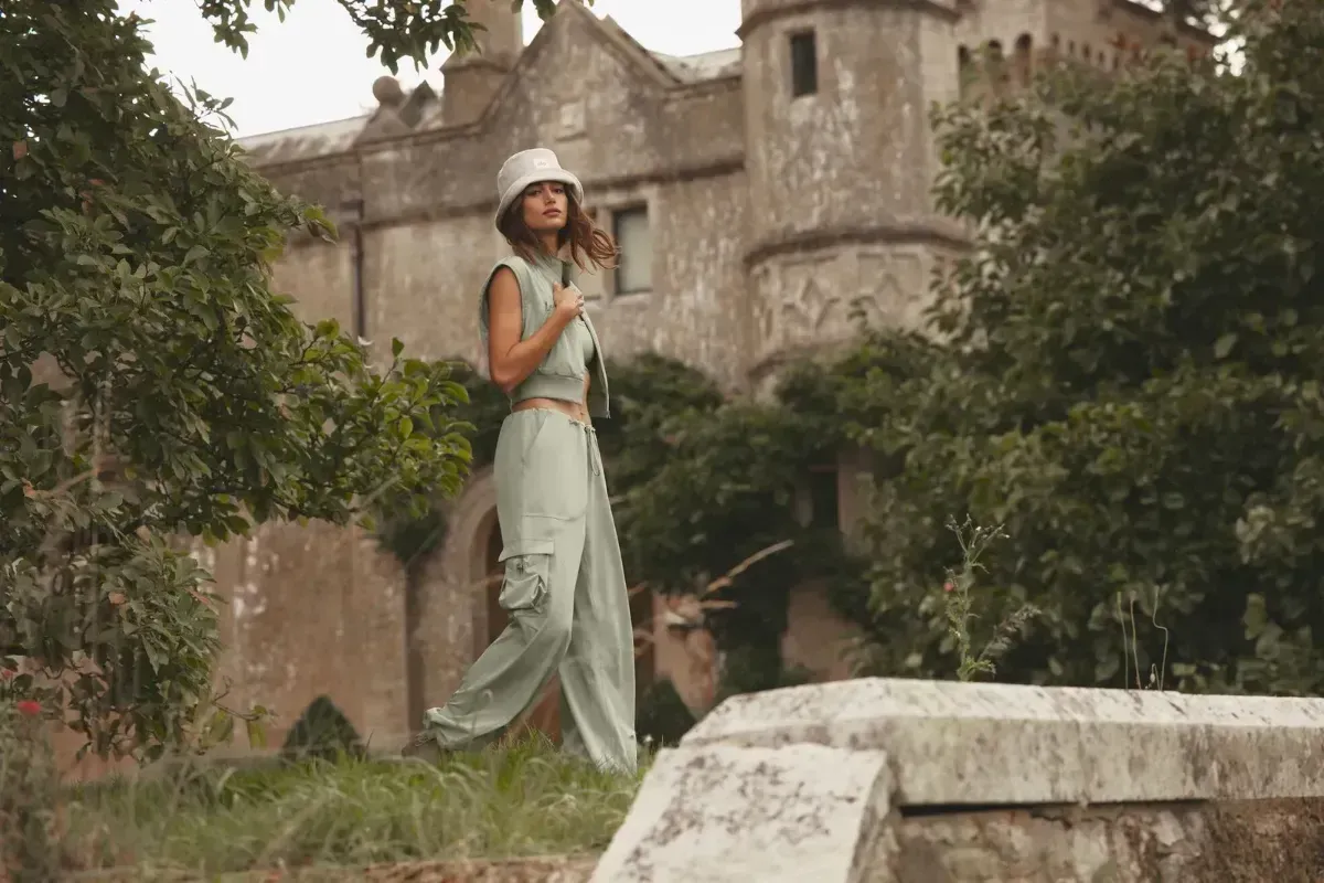 Woman in a light green outfit stands in front of an old stone building and lush trees.