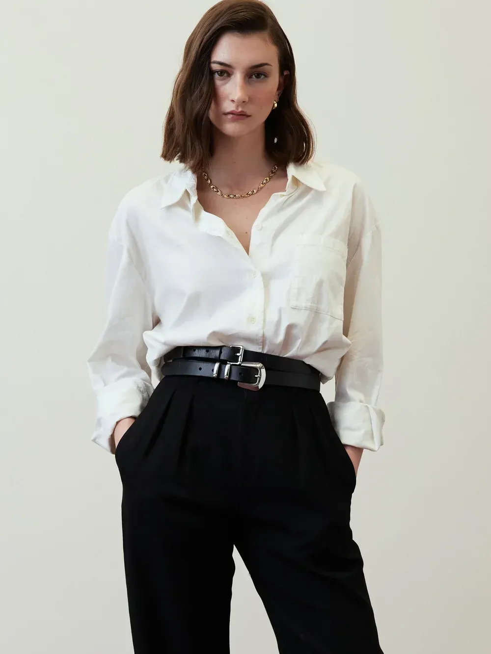 Person in a white button-down shirt and black trousers, standing against a plain light wall.