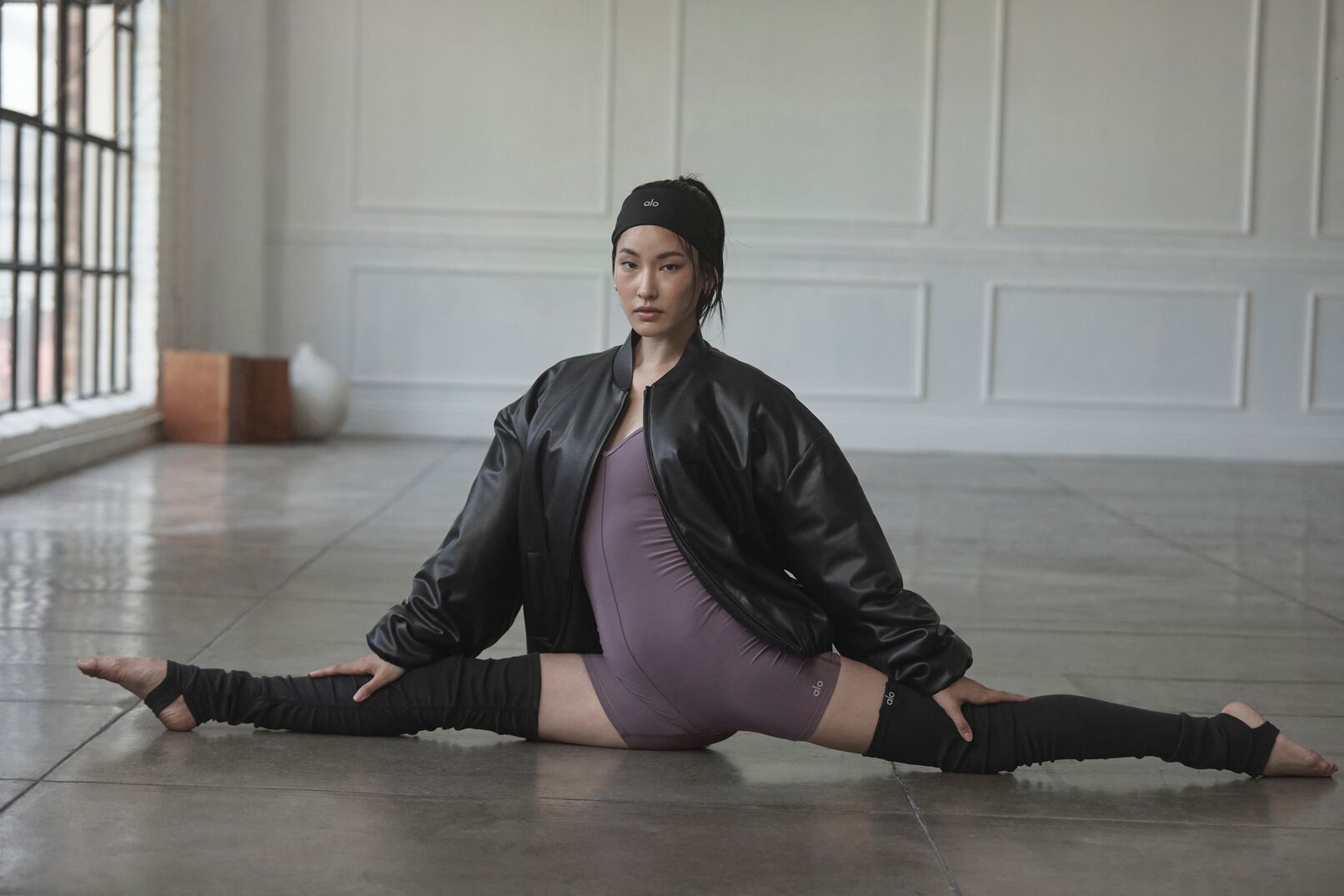 Dancer in a side split on a studio floor, wearing a black jacket and purple leotard.
