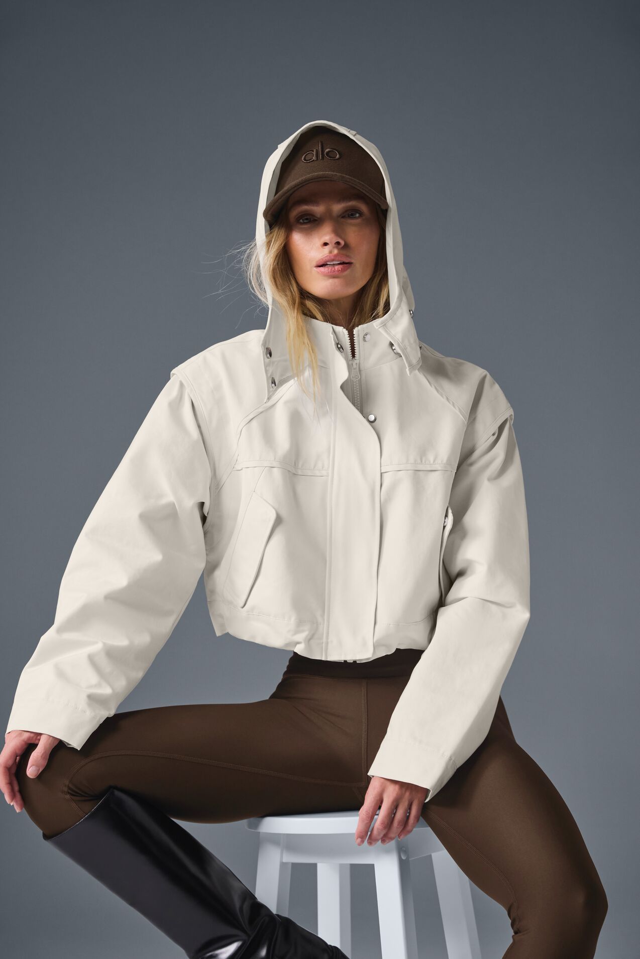 Model in a cream hooded jacket and brown leggings, posing seated on a white stool against a gray background.