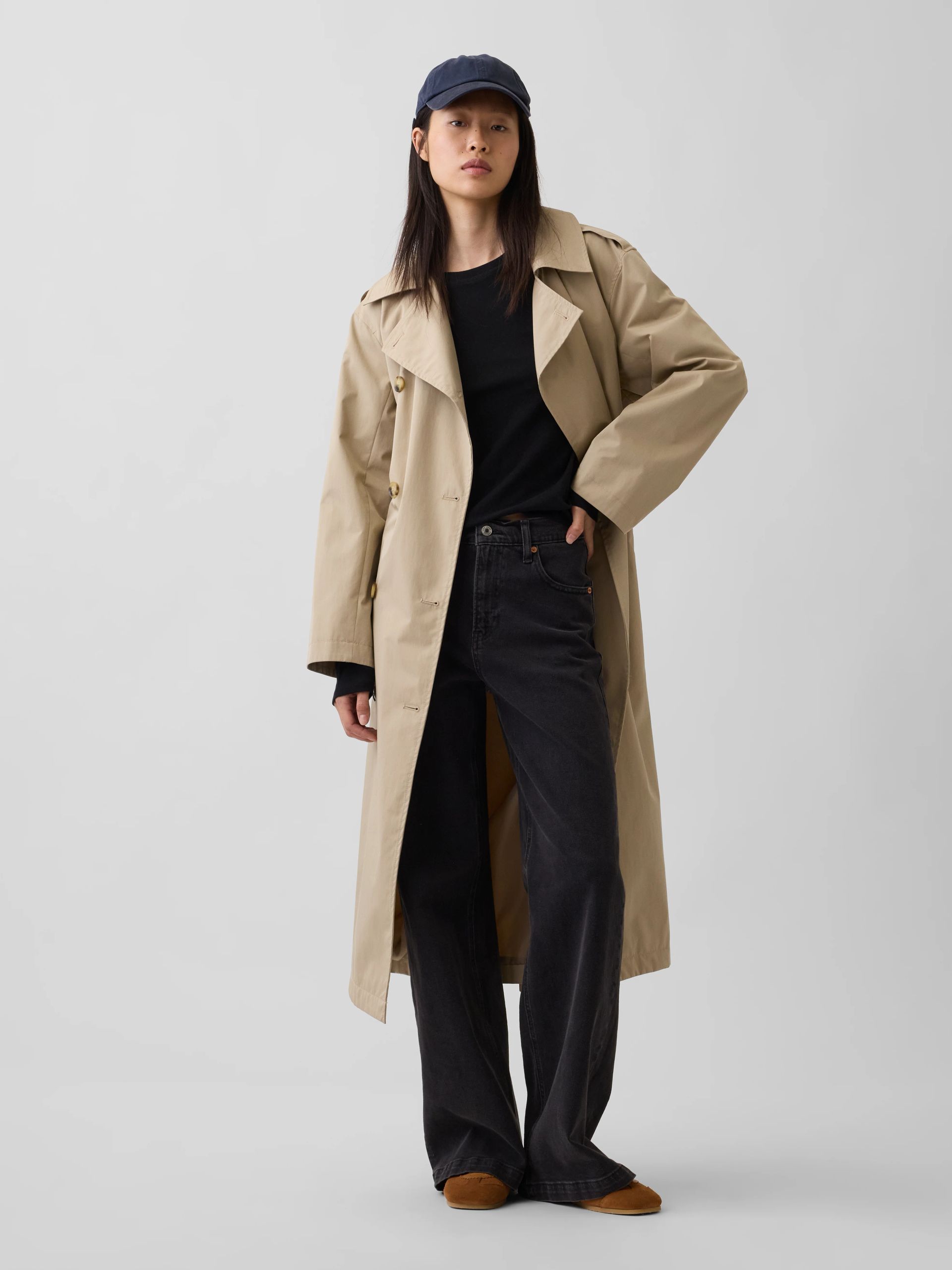 Person in a beige trench coat, black shirt, dark jeans, and cap standing against a plain background.