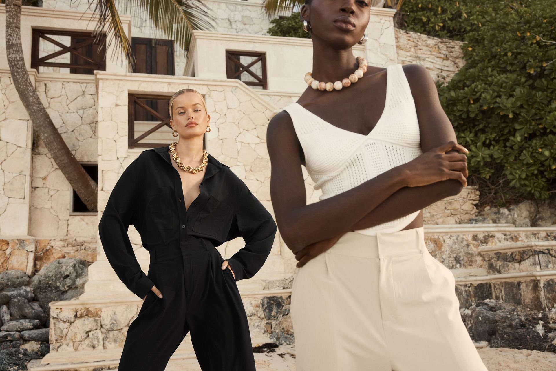 Two models pose outdoors in neutral-toned outfits on stone steps by a rustic building.