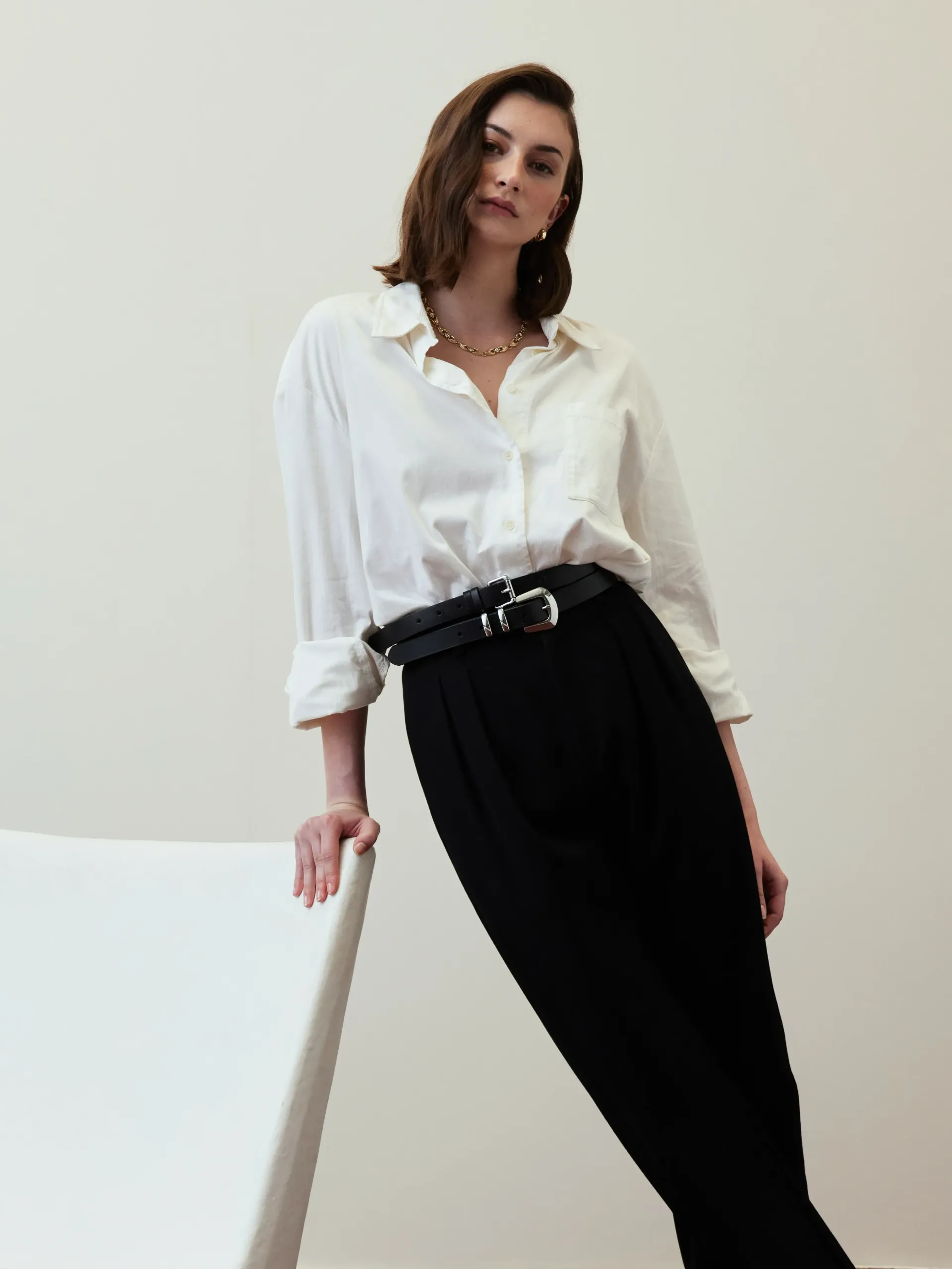 Woman in a white blouse and black pants leaning against a chair in a minimalist room
