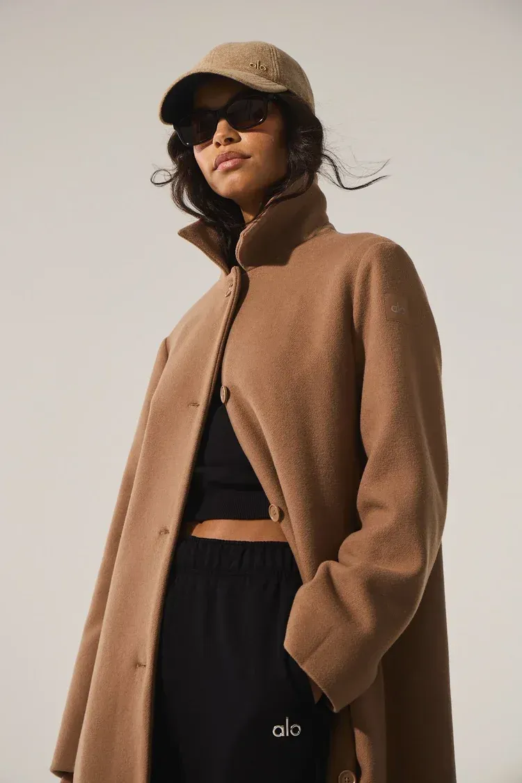 Woman in brown jacket and black pants wearing sunglasses and a cap against a plain background