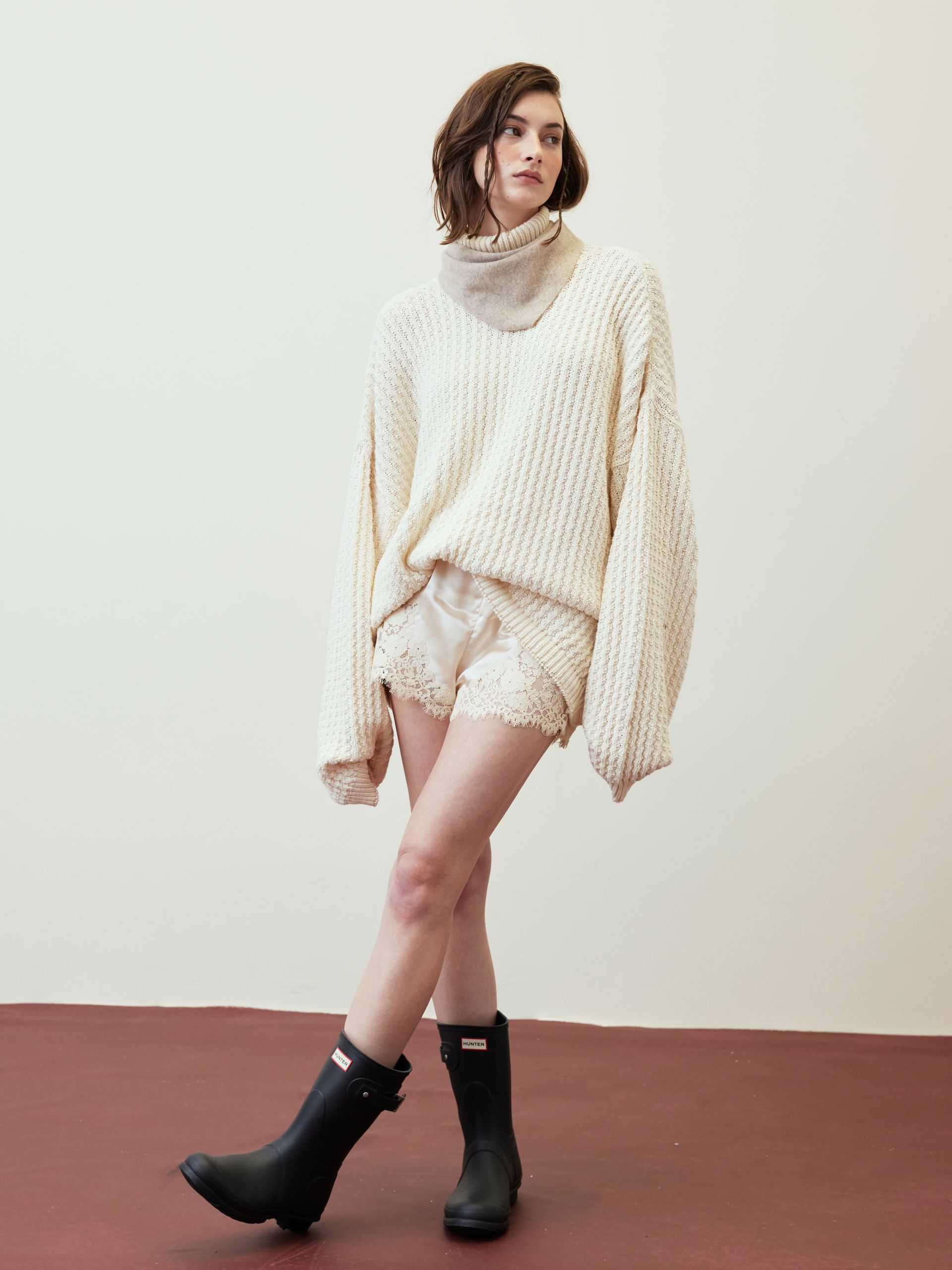 Model in cream knit sweater and shorts with black ankle boots, posing against a pale backdrop.