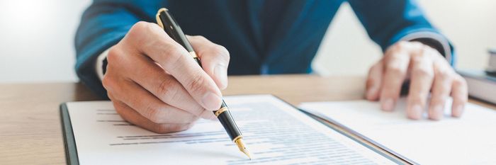 La mano di una persona che tiene una penna mentre scrive un documento su una scrivania di legno.