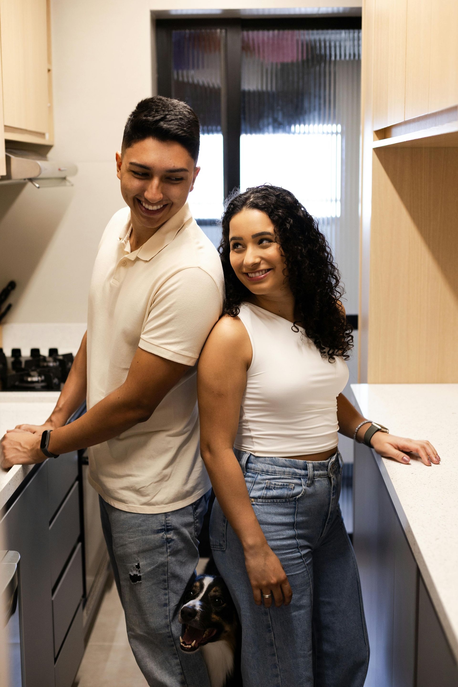 Couple smiling, back to back, in a modern kitchen. Man in beige shirt, woman in white tank top, dog visible.