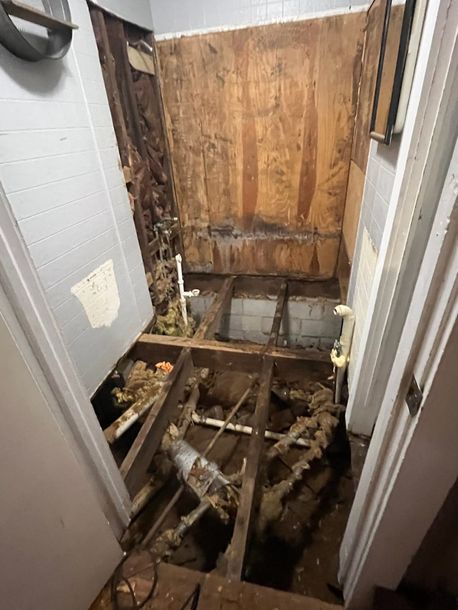 Collapsed bathroom floor with exposed joists and debris, showing extensive water damage and rot
