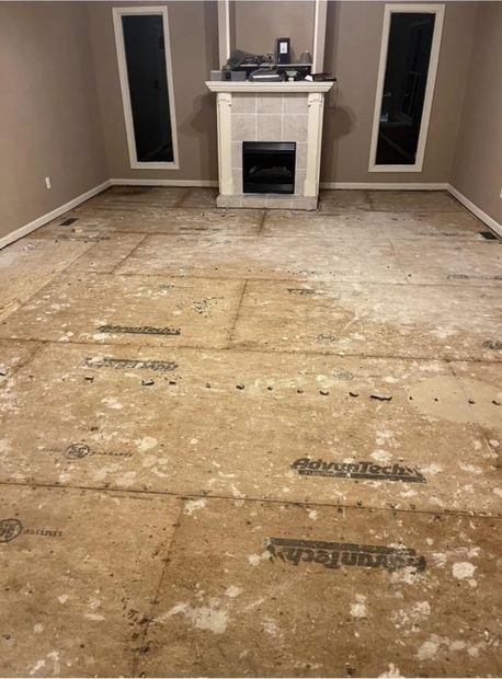Empty room with removed carpet, exposed subfloor, and a white fireplace centered between two windows.